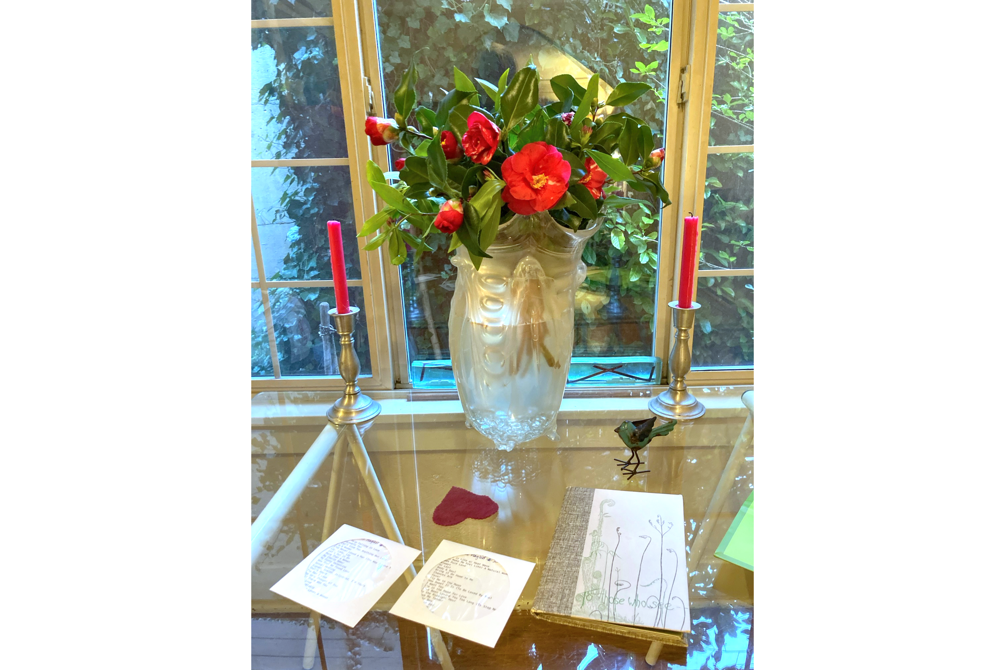 Photo of vase of red camellias on a class table with two candles, a heart, 2 cds, and a book.