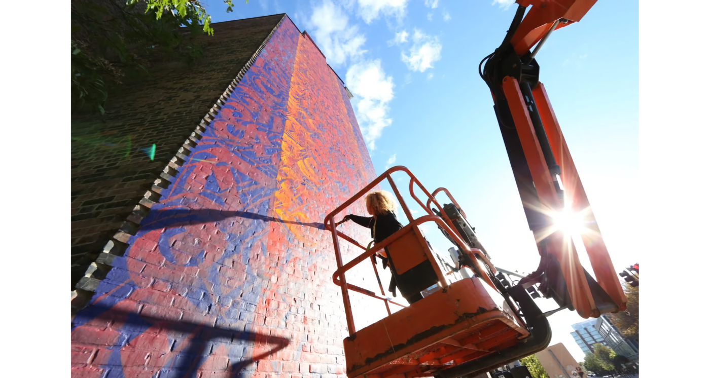 Photo of woman on boom lift painting side of building in bright colors
