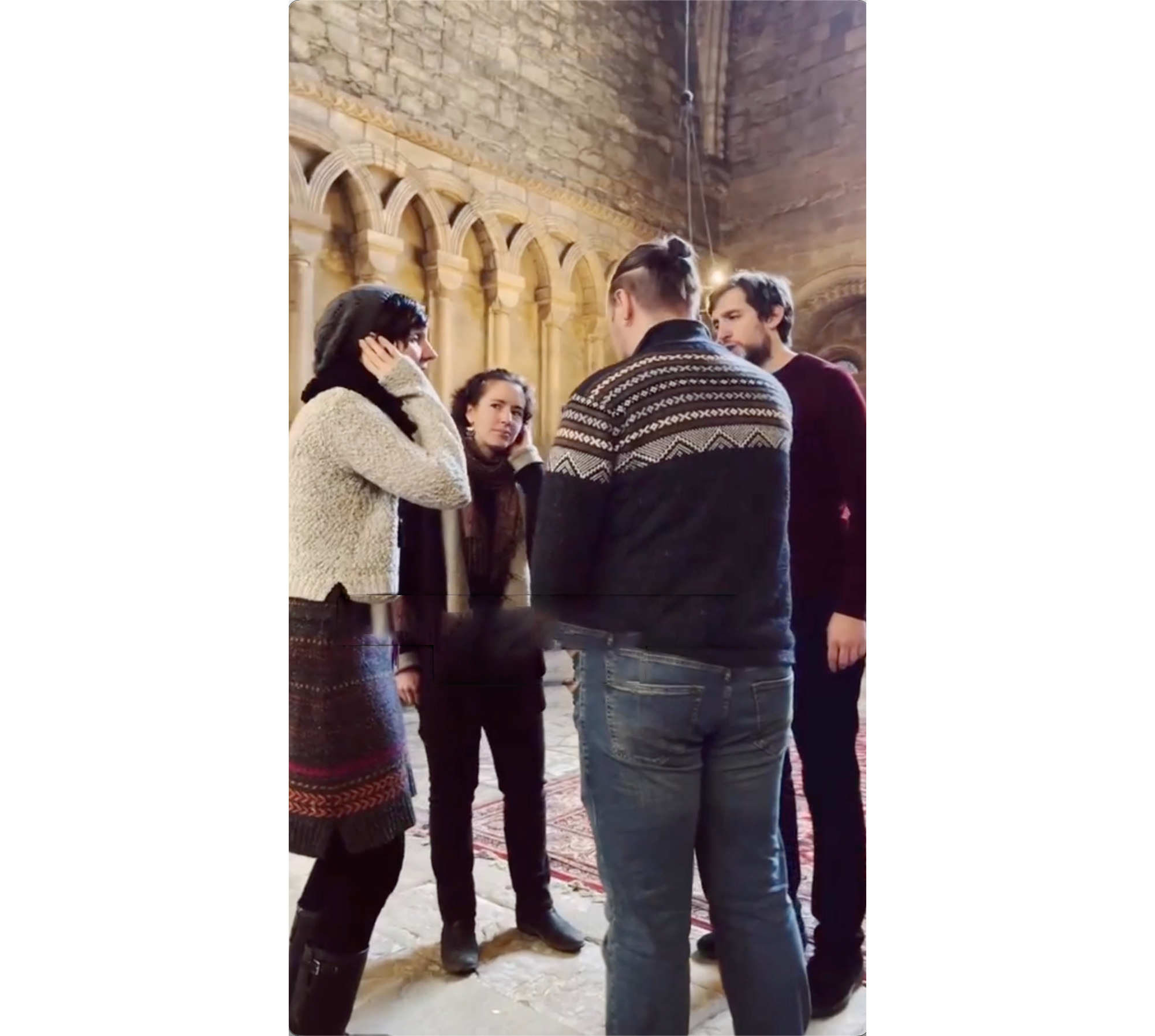 Photo of two female and two male singers, standing in a close circle, each with one hand cupped to their ear, with arched cathedral wall in background