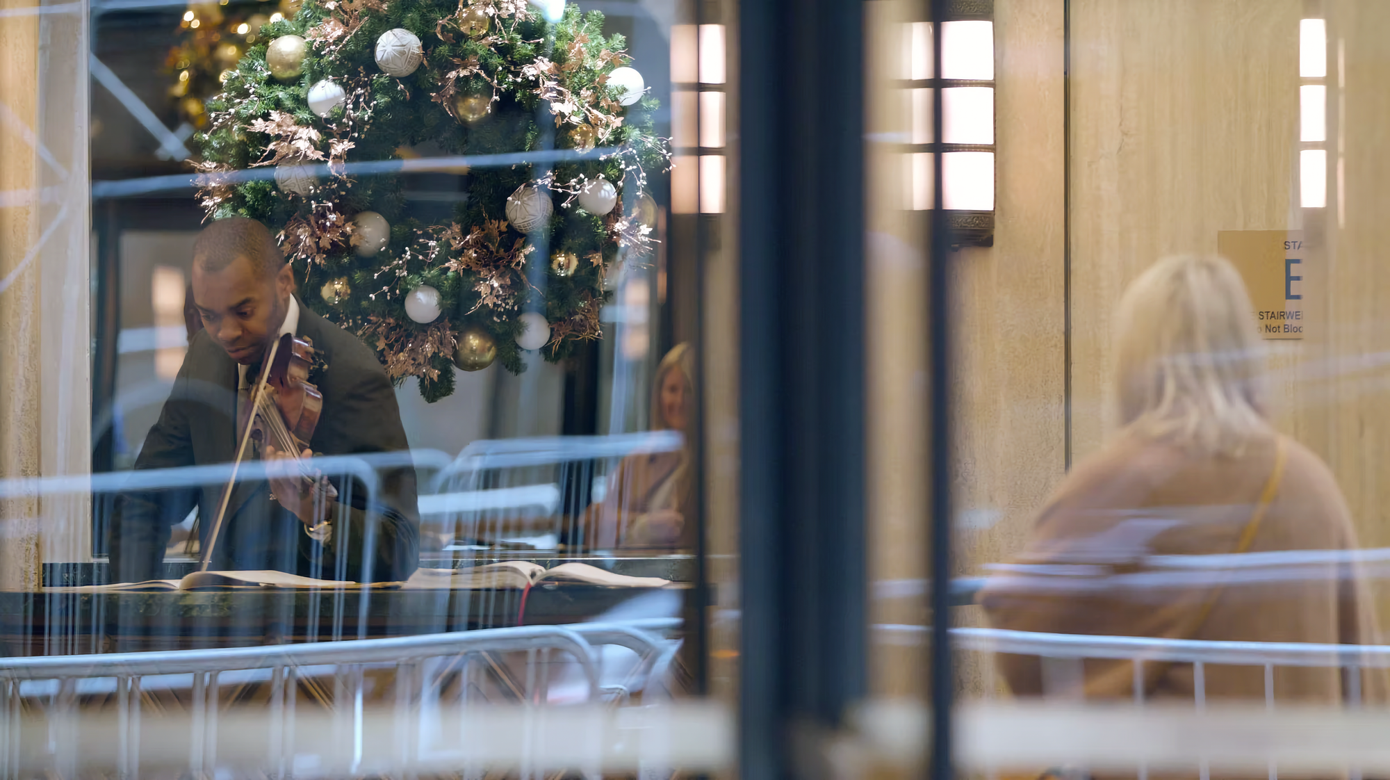 Photo of Yves Deshommes looking down at sheet music while playing violin at his desk, with a holiday wreath in foreground, and a seated woman in a coat to the right listening.