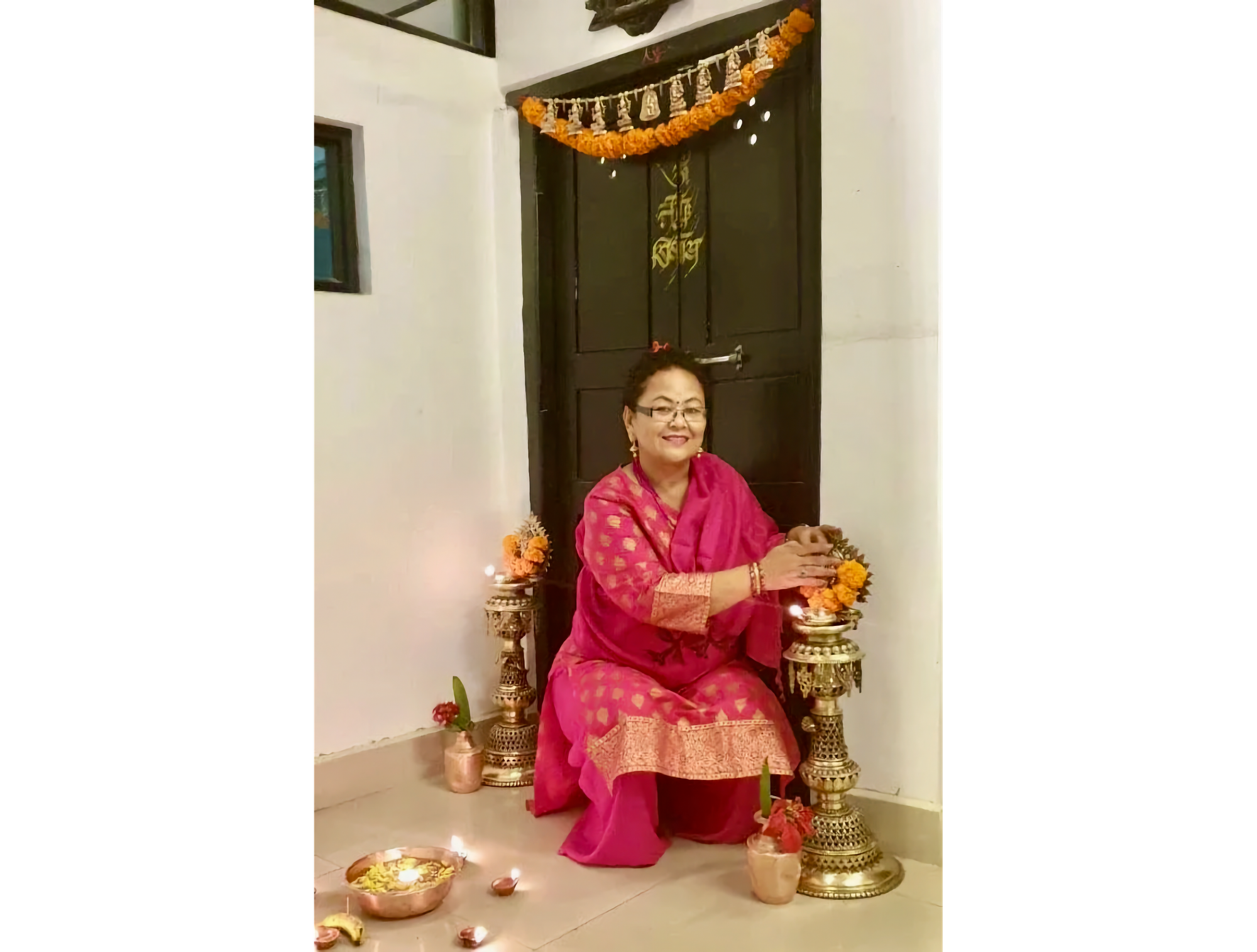 Photo of Nepali woman in red traditional cress seated in front of door with her hands on flowers atop gold stand