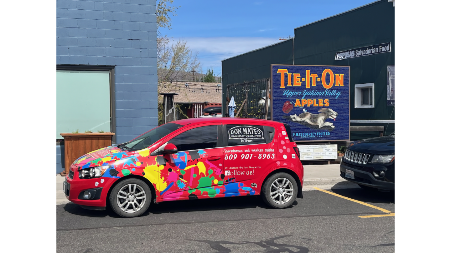 A red compact car with a colorful paint-splatter wrap advertising Don Mateo SalvaMex Restaurant, parked in front of a vintage mosaic sign for an apple producer.