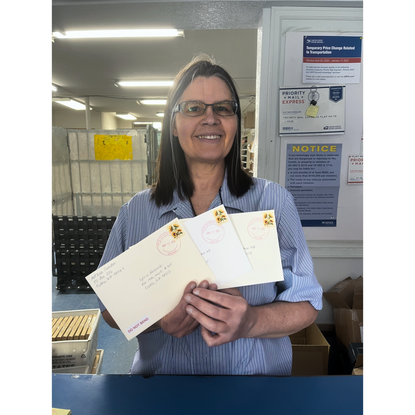 Photo of woman postal worker wearing glasses and holding 3 stamped envelopes.