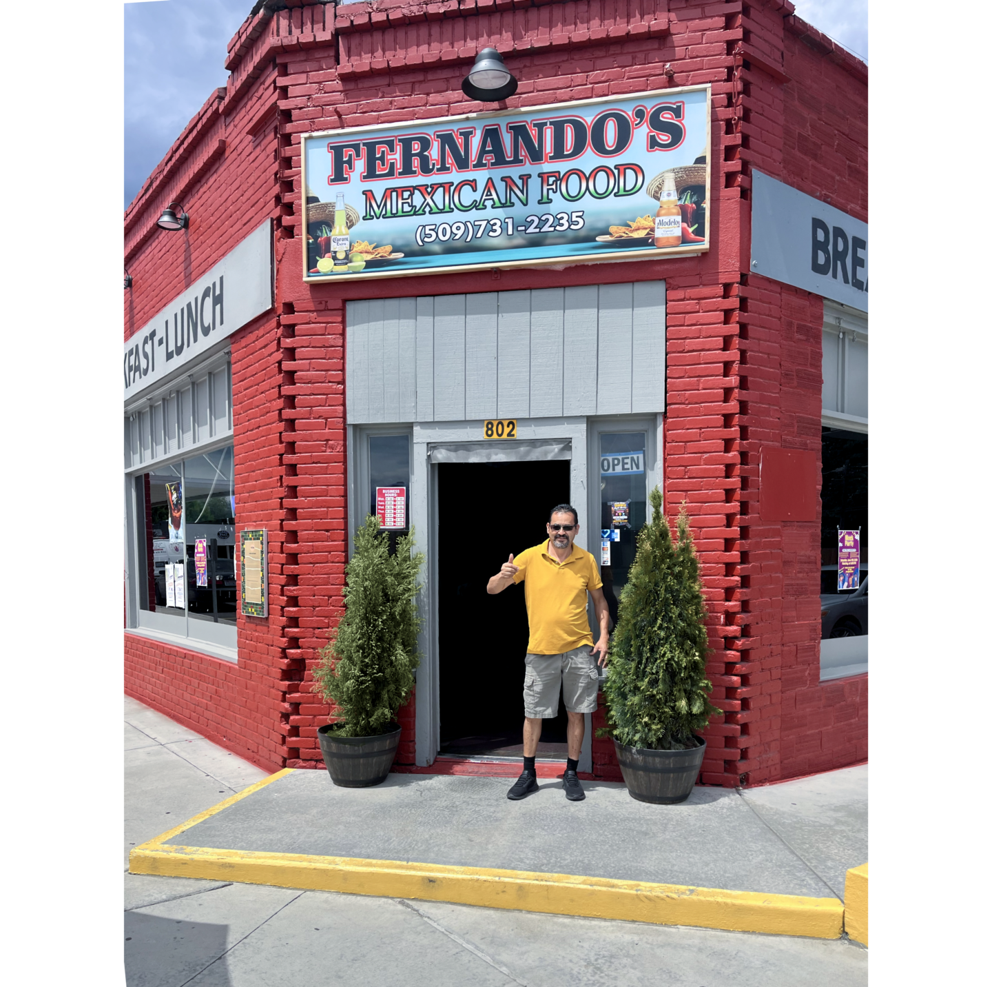 Photo of Fernando's restaurant, single story red brick building, with Fernando in front in grey shorts and yellow shirt holding right thumb up and wearing sunglasses, in front of open door.