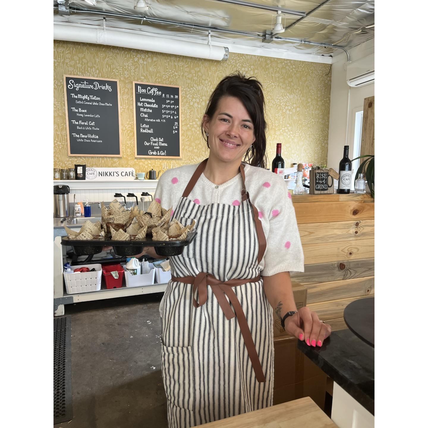 Photo of wman wearing striped apron over white shirt with pink polka dots, holding tray of pastries, with price boards in background.
