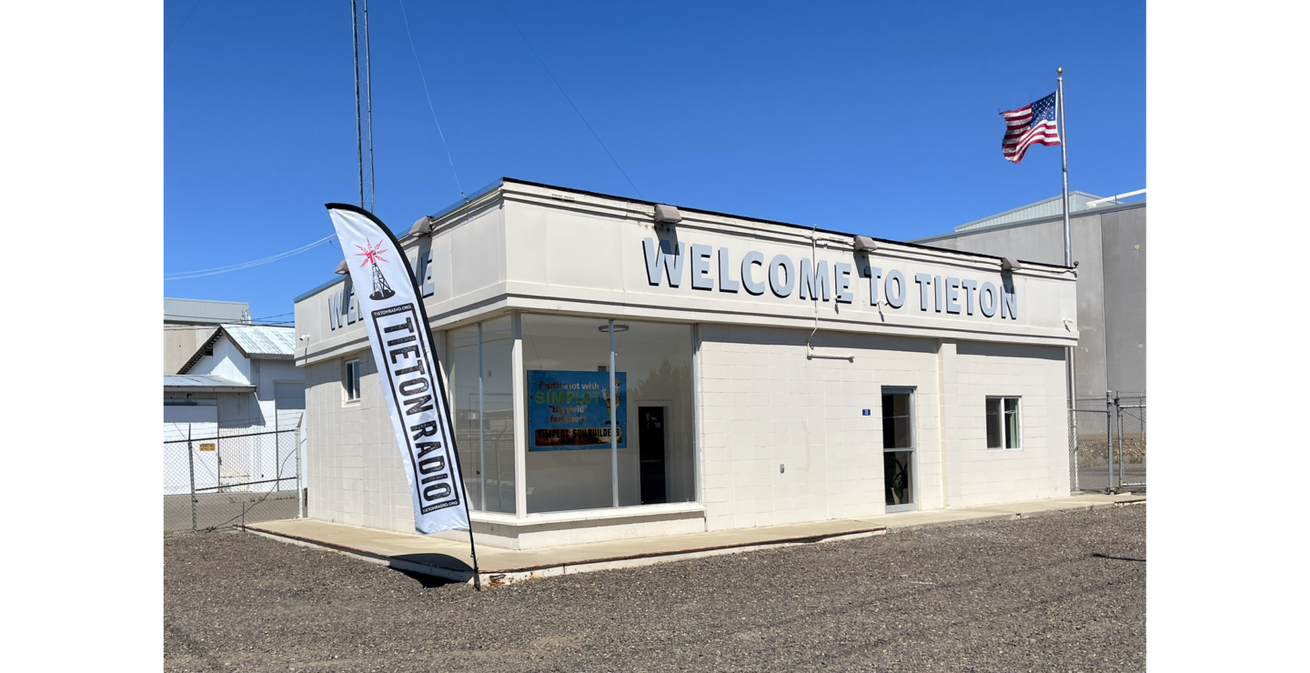 Photo of small white building with sign "Welcome to Tieton" and Tieton Radio banner on left and American flag on right
