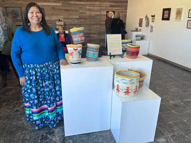 Wahpeniat stands in blue top and blue patterned long skirt next to display of her baskets on white pedestals