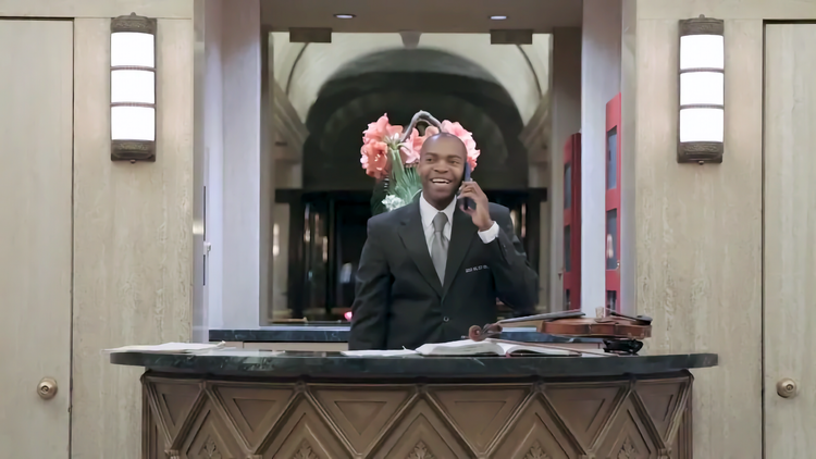 Photo of black man in suite on phone behind lobby desk, in front of mirror, with violin on desk to his right