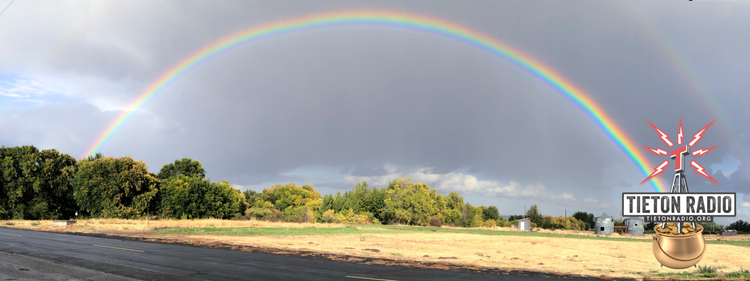 Photo montage of rainbow over trees and brown field with radio station logo in pot of gold at right end.