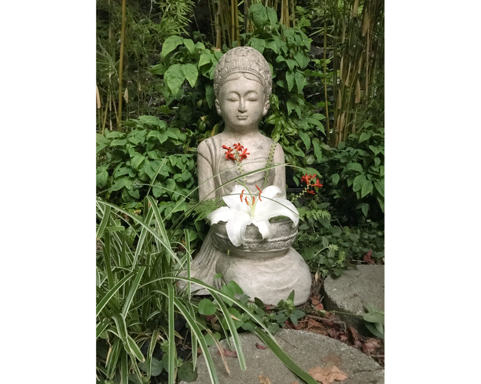 Photo of concrete cast sculpture of Buddhist goddess holding white lily in her hands in front, with ivy and fence in background