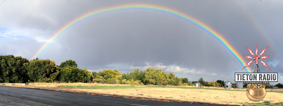 Photo montage of rainbow over trees and brown field with radio station logo in pot of gold at right end.