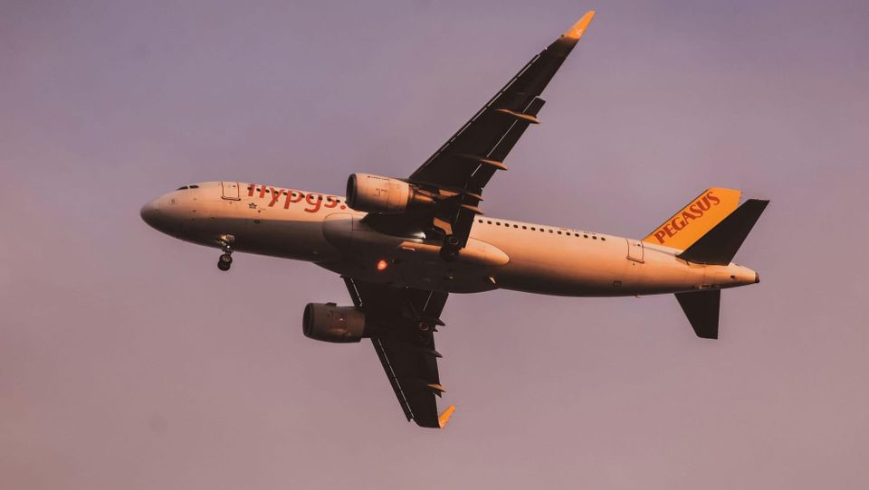 Photo of a modern airplane flying low over green fields with mountains in background and partly cloudy sky
