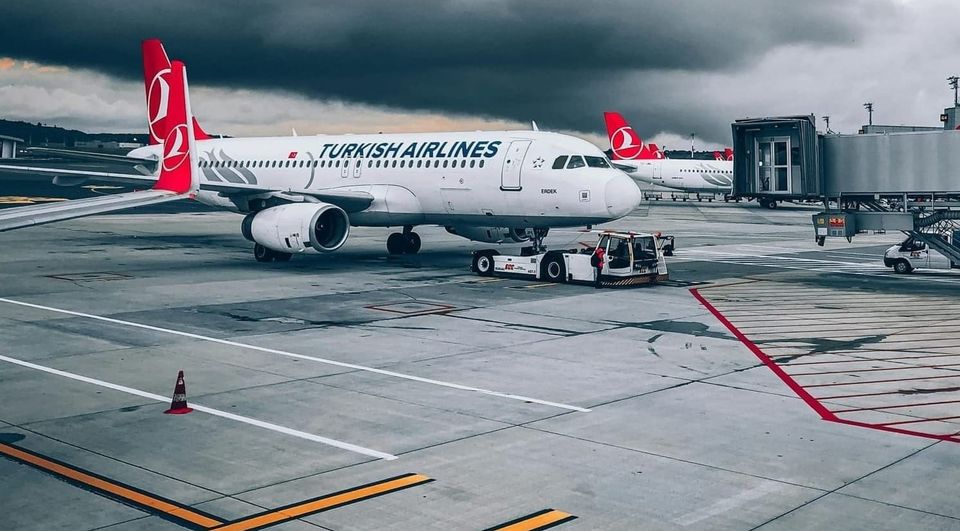 Photo of a modern airplane flying low over green fields with mountains in background and partly cloudy sky