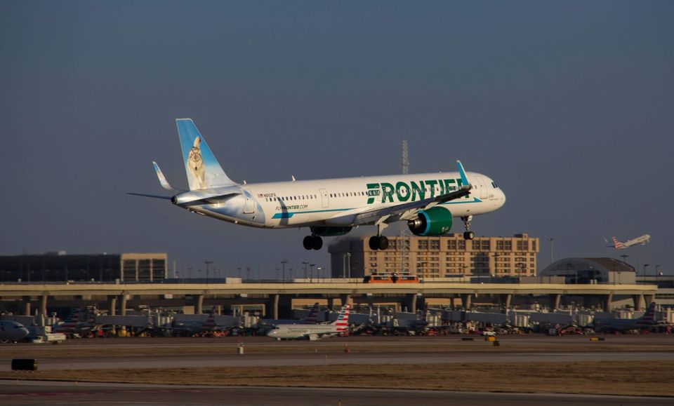 Photo of a modern airplane flying low over green fields with mountains in background and partly cloudy sky
