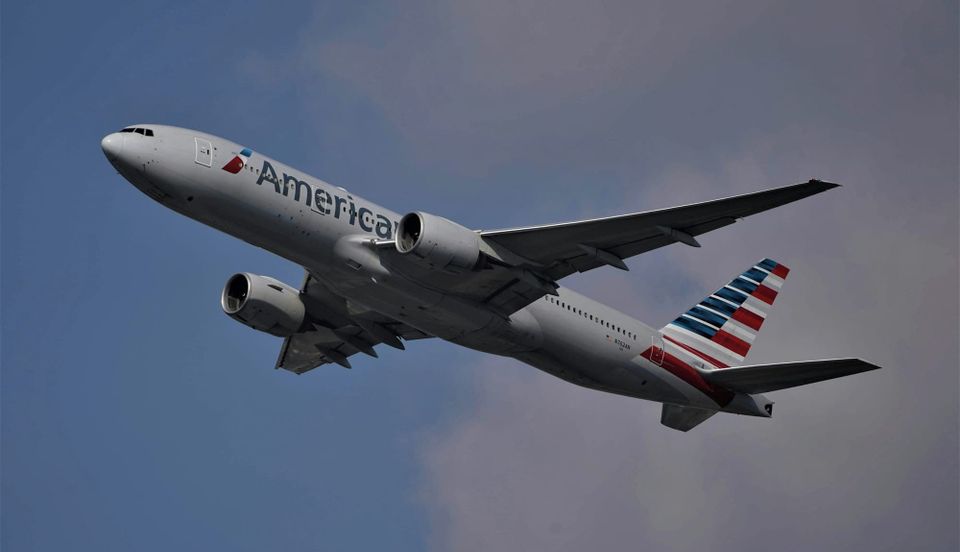 Photo of a modern airplane flying low over green fields with mountains in background and partly cloudy sky