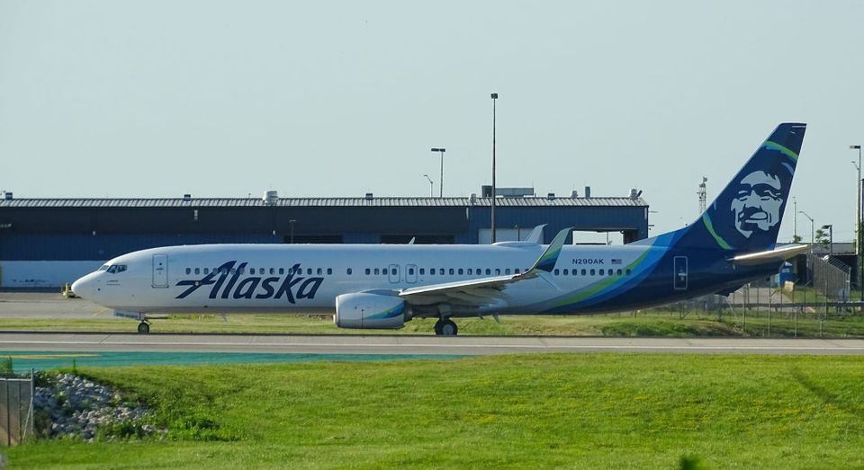 Photo of a modern airplane flying low over green fields with mountains in background and partly cloudy sky