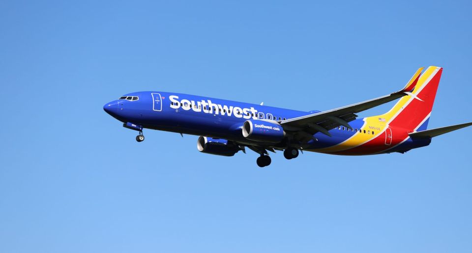 Photo of a modern airplane flying low over green fields with mountains in background and partly cloudy sky