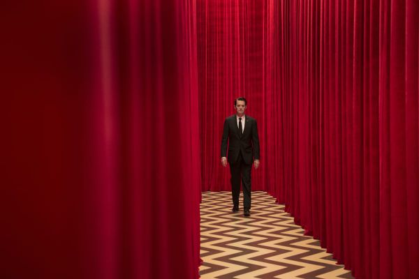 A man walks down a corridor created by red curtains, and the floor is a zigzag pattern of black and white. it is an image from the Red Room in Twin Peaks. 