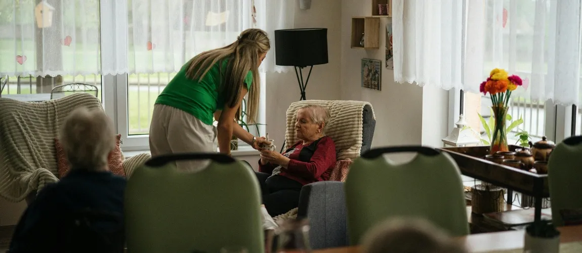 Eine Frau reicht einer Seniorin, die im Sessel sitzt, eine Tasse.