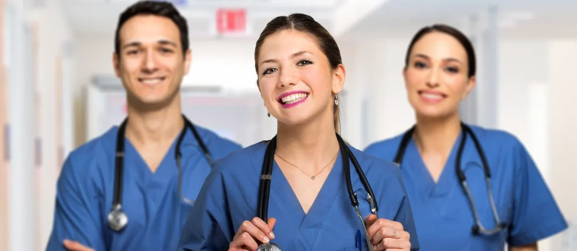 Drei Pflegepersonen in blauer Kleidung lächeln in die Kamera.