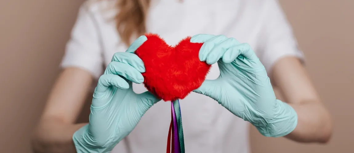 Eine Frau mit einem weißen Oberteil und Handschuhen hält ein Herz aus Plüsch in die Kamera.
