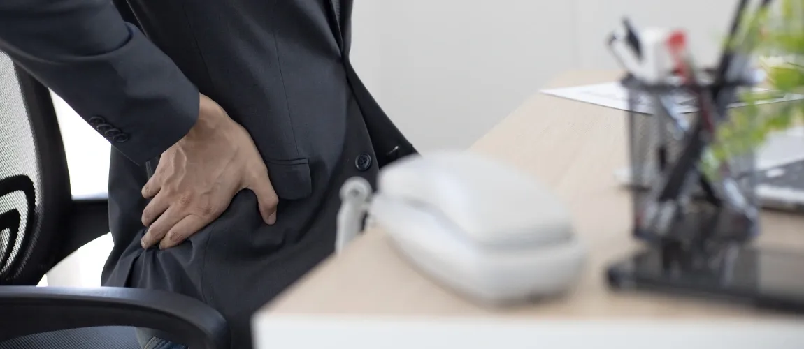 Eine Person sitzt am Schreibtisch und stützt sich mit einer Hand den Rücken.