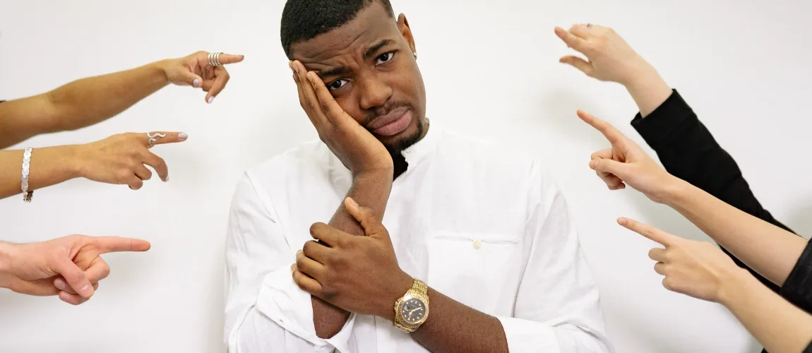 Ein Mann in einem weißen Oberteil schaut belastet in die Kamera, von links und rechts zeigen Finger auf ihn. 