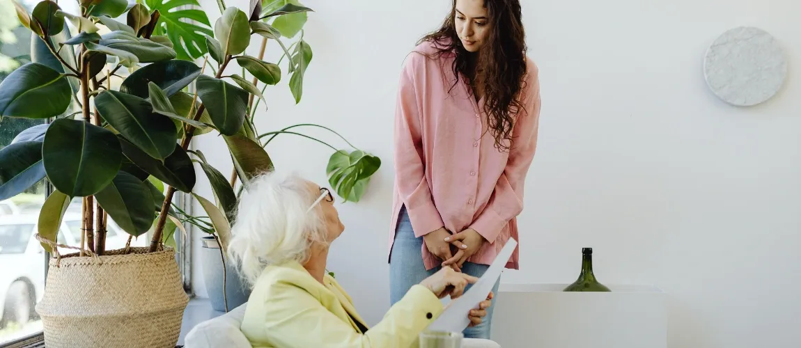 Eine Seniorin sitzt auf einem Sessel und zeigt auf ein Blatt Papier, daneben steht eine junge Frau.
