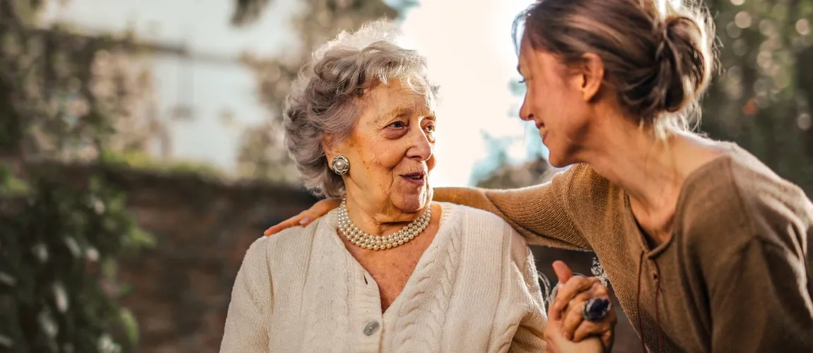 Eine Frau legt einen Arm um eine Seniorin und lächelt.