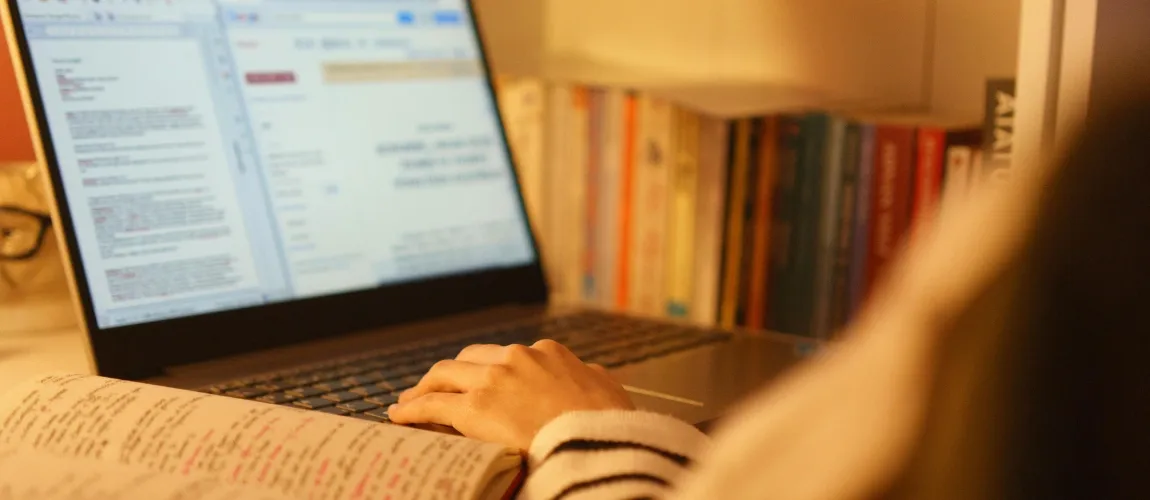 Eine Person sitzt vor einem Laptop, daneben liegt ein aufgeschlagenes Notizbuch. 
