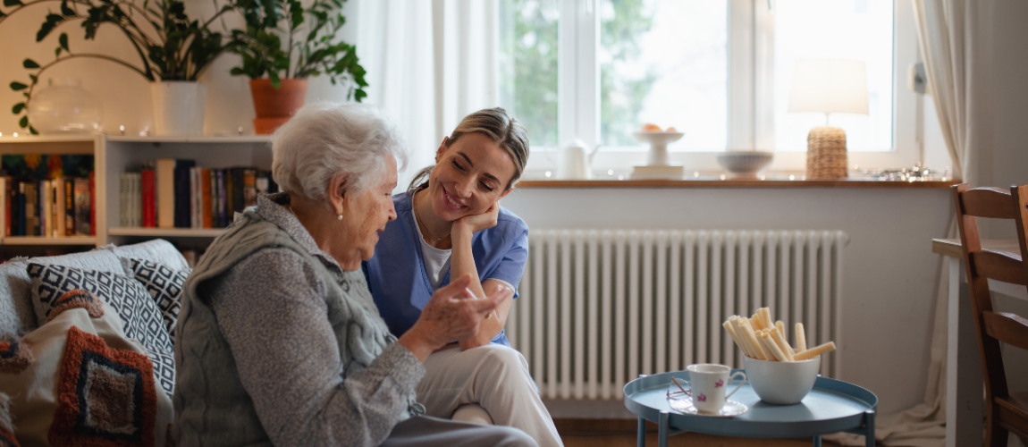 Eine Pflegehelferin sitzt mit einer Patientin auf dem Sofa.