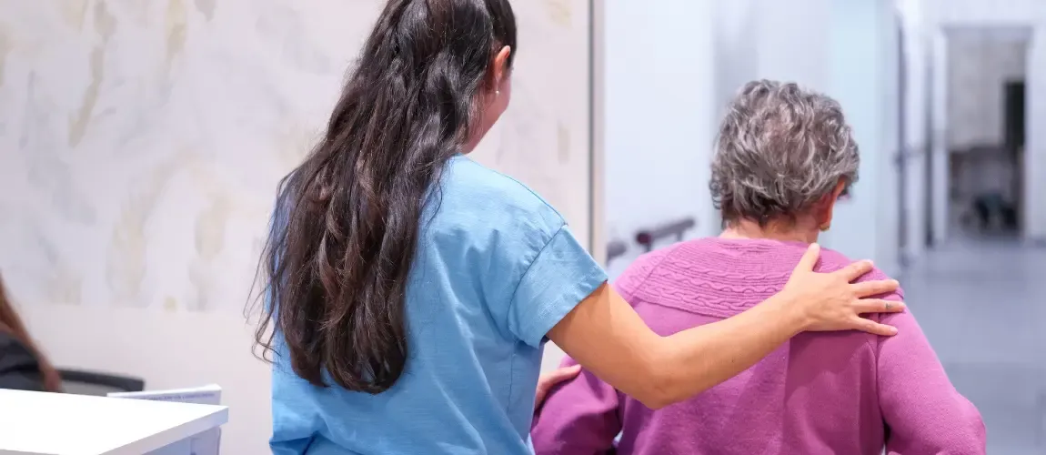 Pflegekraft hält Patientin an der Schulter und führt sie durch den Gang.