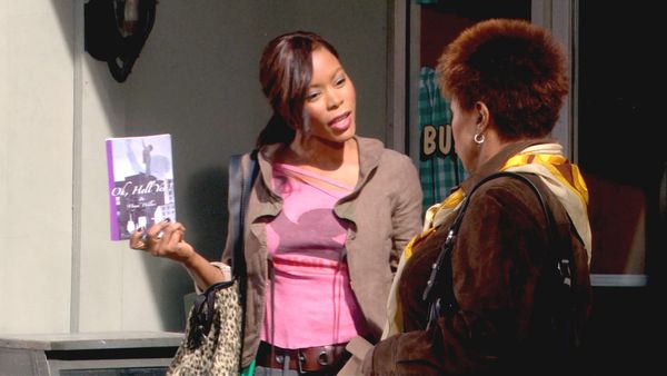 Maya holds up her book Oh, Hell Yes!, wearing a brown jacket and pink shirt
