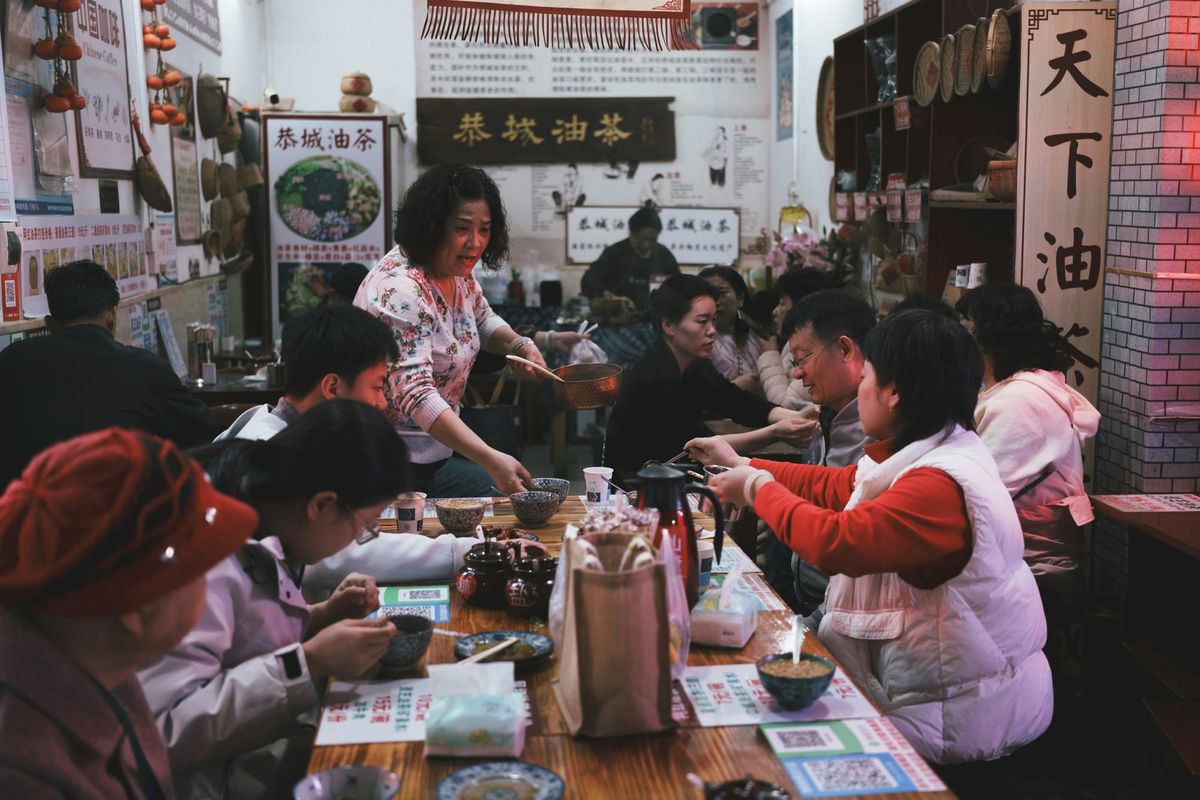 Locals sitting closely at a long wooden table in a busy regional Chinese restaurant while a server pours tea into small bowls