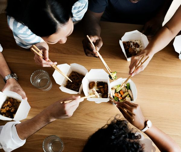 A group of friends eating Western-style Chinese food from classic white paper boxes using chopsticks