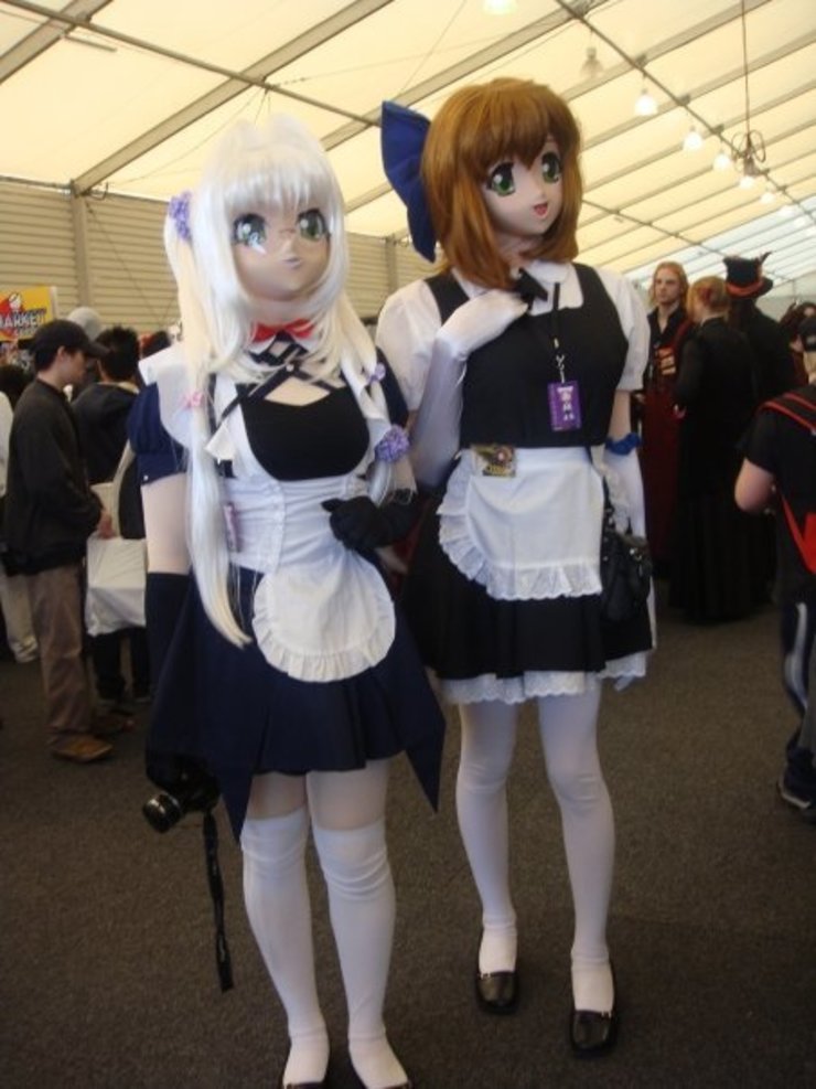 two people wearing fully enclosing anime mask heads with fixed, blankly happy expressions.