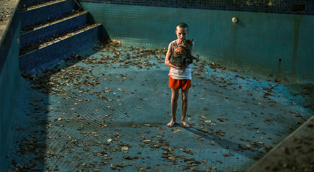 A small child holding a cat stands in a dirty, empty backyard pool.