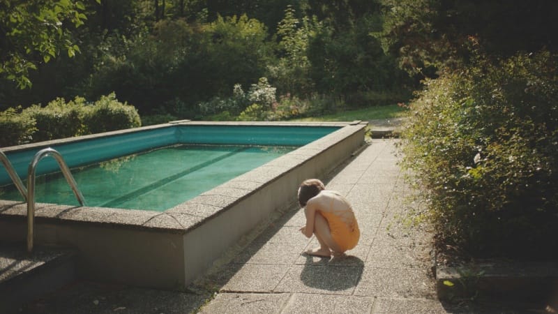 A small child crouches near a clear, still backyard pool.