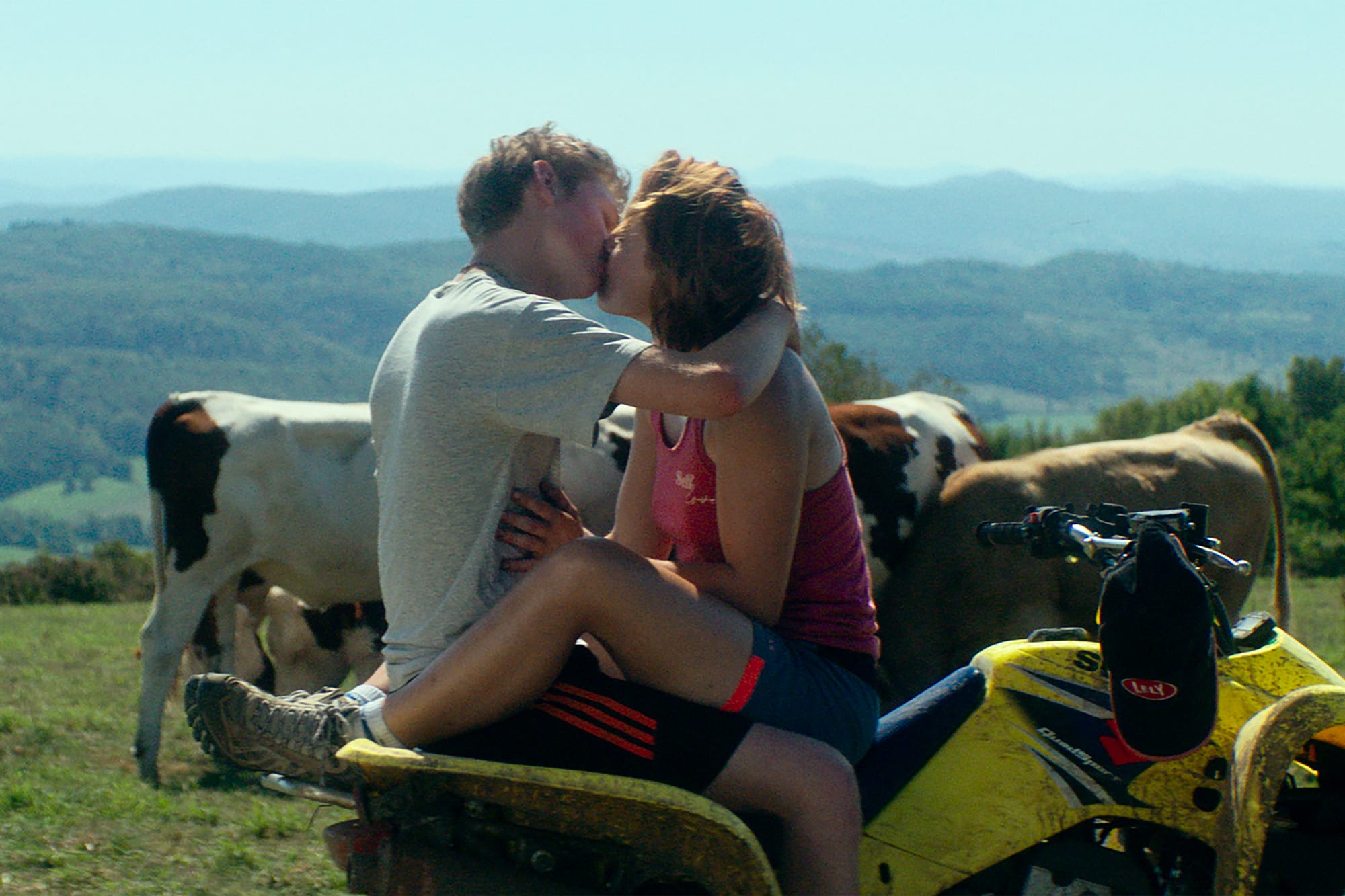 A young man and a young girl sitting face-to-face on a quad bike in a field, kissing. There are cows grazing right behind them, and green hills in the horizon.