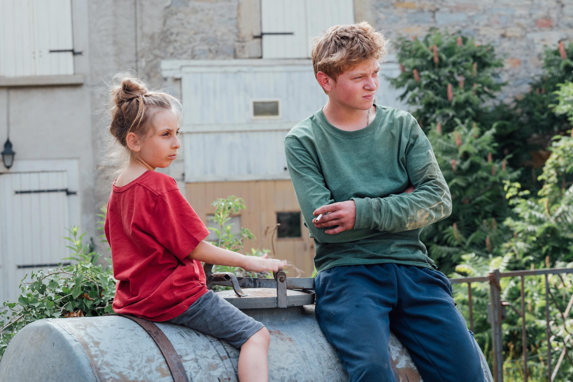A young girl in a red t-shirt and an older boy in a green shirt, both sitting on a milk container outside, looking to the right.