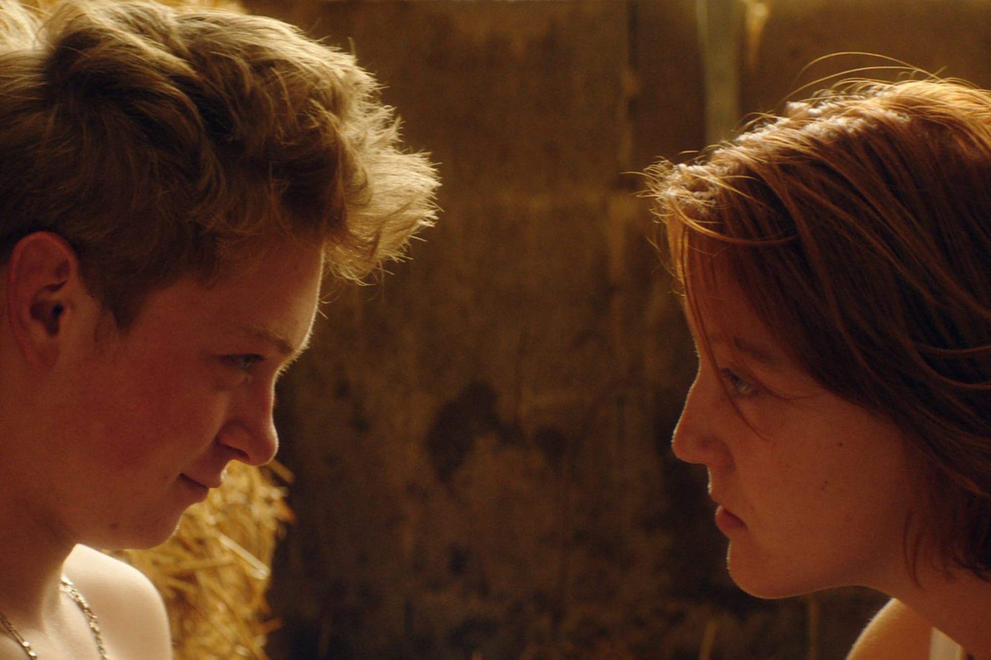 A close-up on the profiles of a young boy and a young girl looking at each other. They're in a stable filled with yellow straw.
