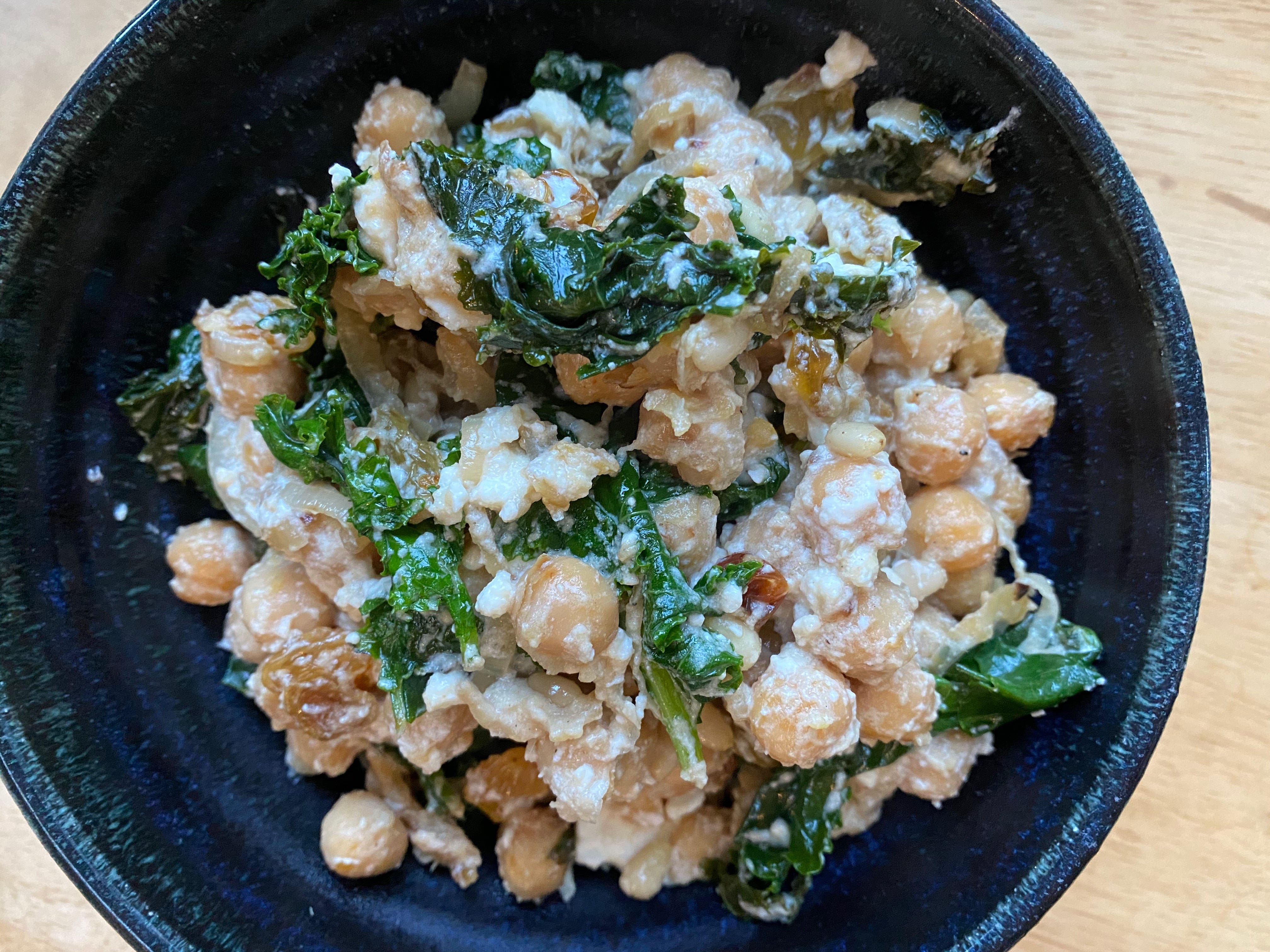 A blue ceramic bowl full of chickpeas, kale, and golden raisins in a creamy tahini sauce.