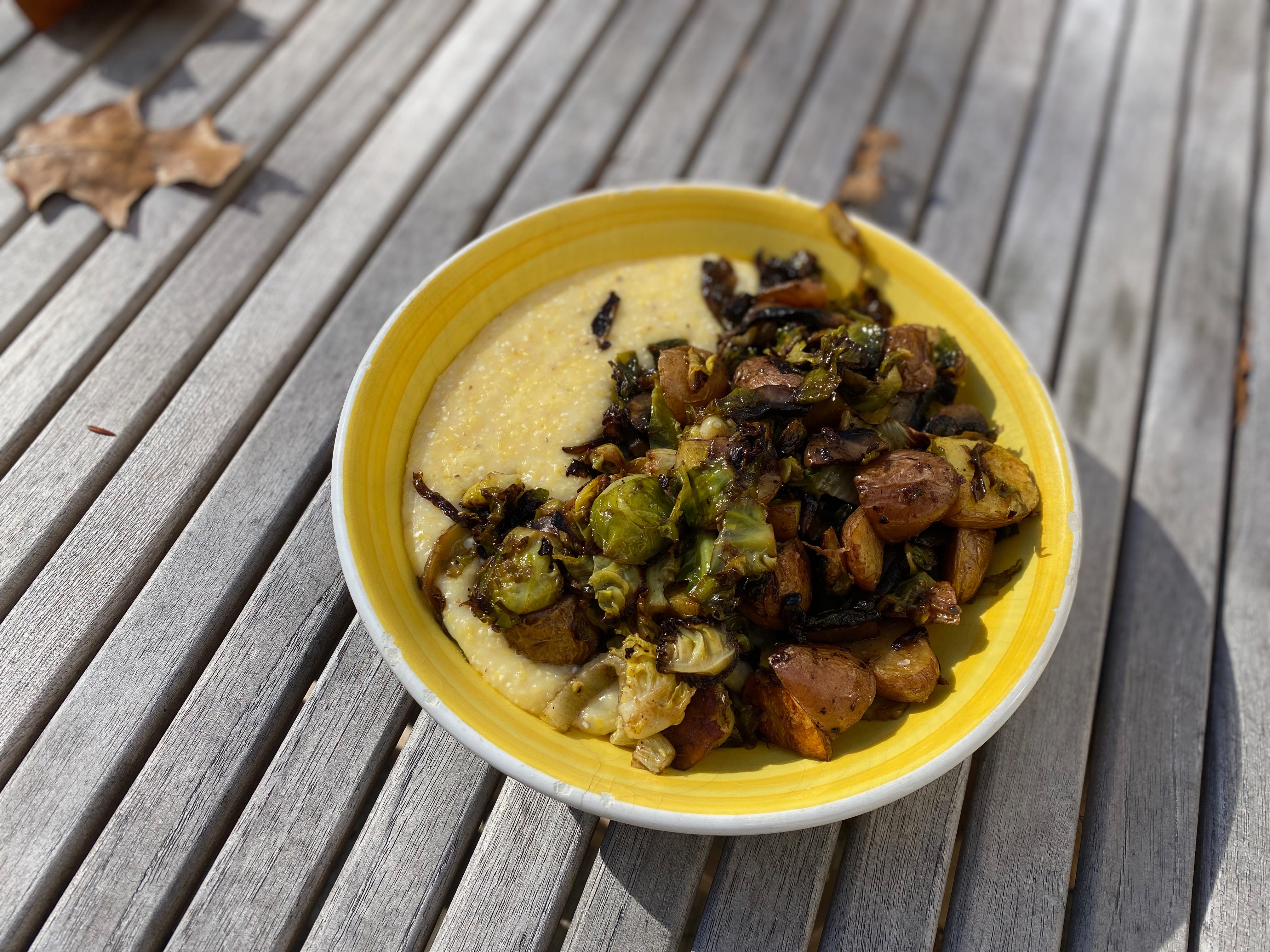 A shallow yellow bowl filled with polenta, Brussels sprouts, and roasted potatoes sits on an outdoor table. 