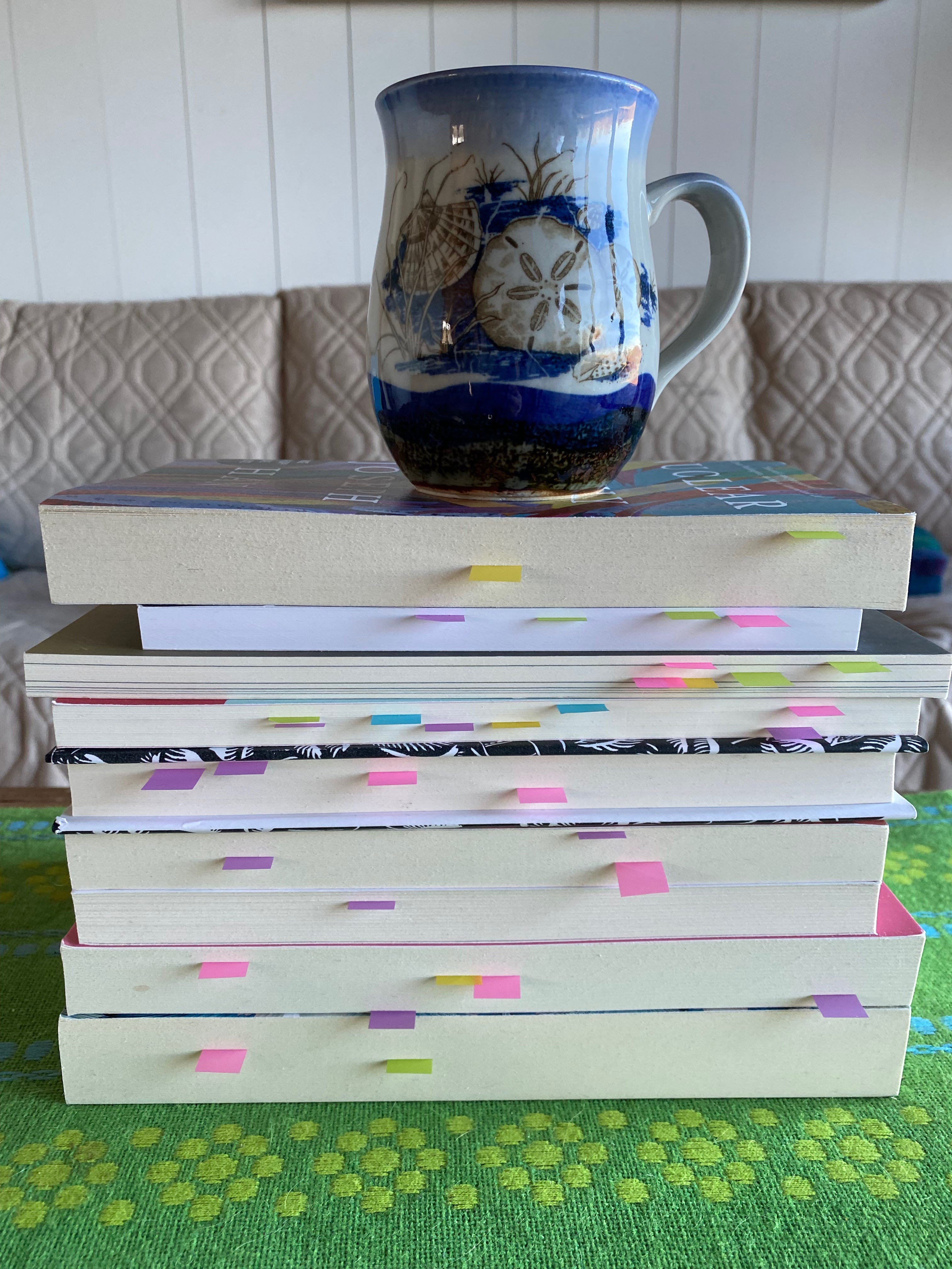 A stack of books, spines facing away. Small colored post-it notes poke out from the pages. A ceramic mug painted with a sand dollar sits on top of the books.