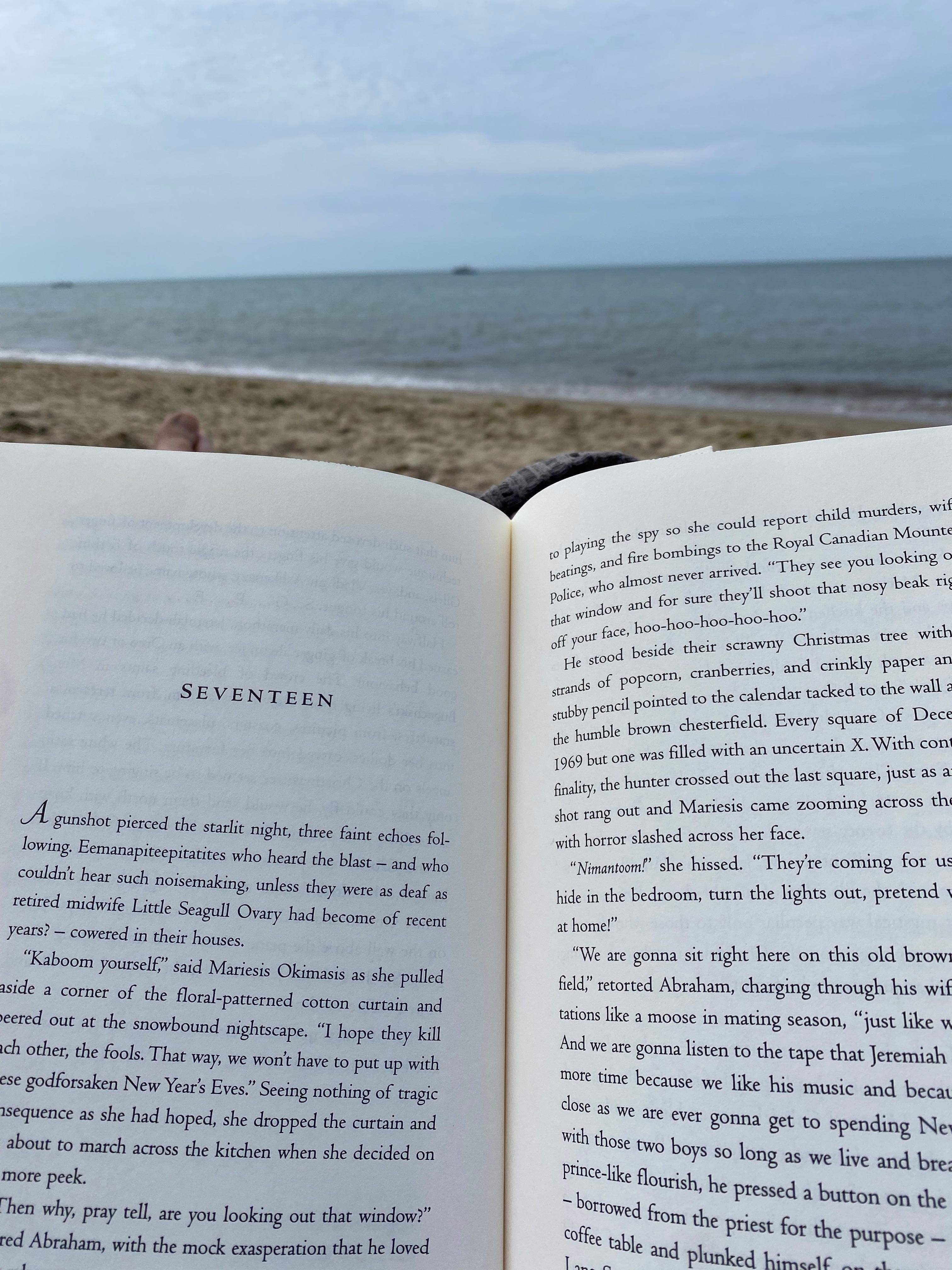 Closeup of an open book propped on my knees; you can see the beach, the ocean, and a cloudy horizon in the background.