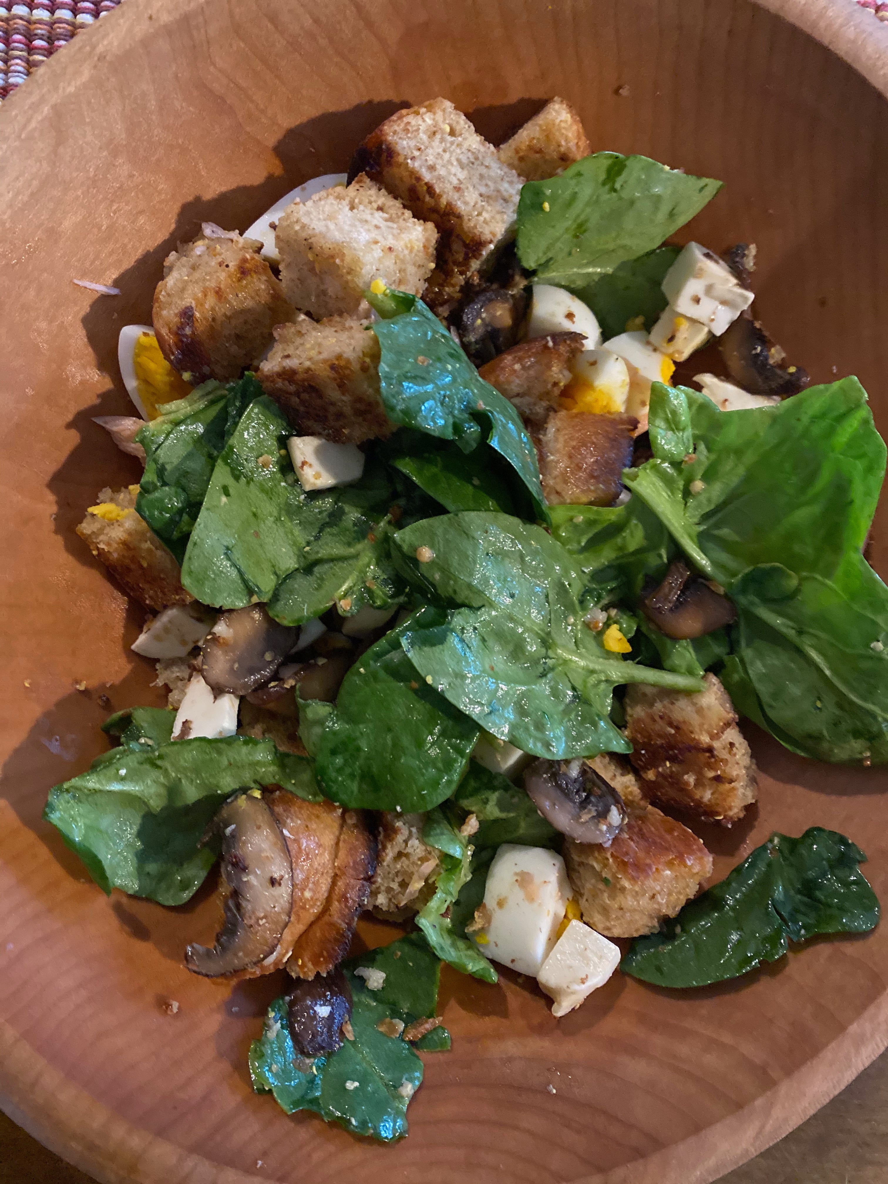 A salad of spinach leaves, chunks of golden brown fried bread, sautéed mushrooms, fresh mozzarella cubes, and hard boiled eggs in a wooden bowl.