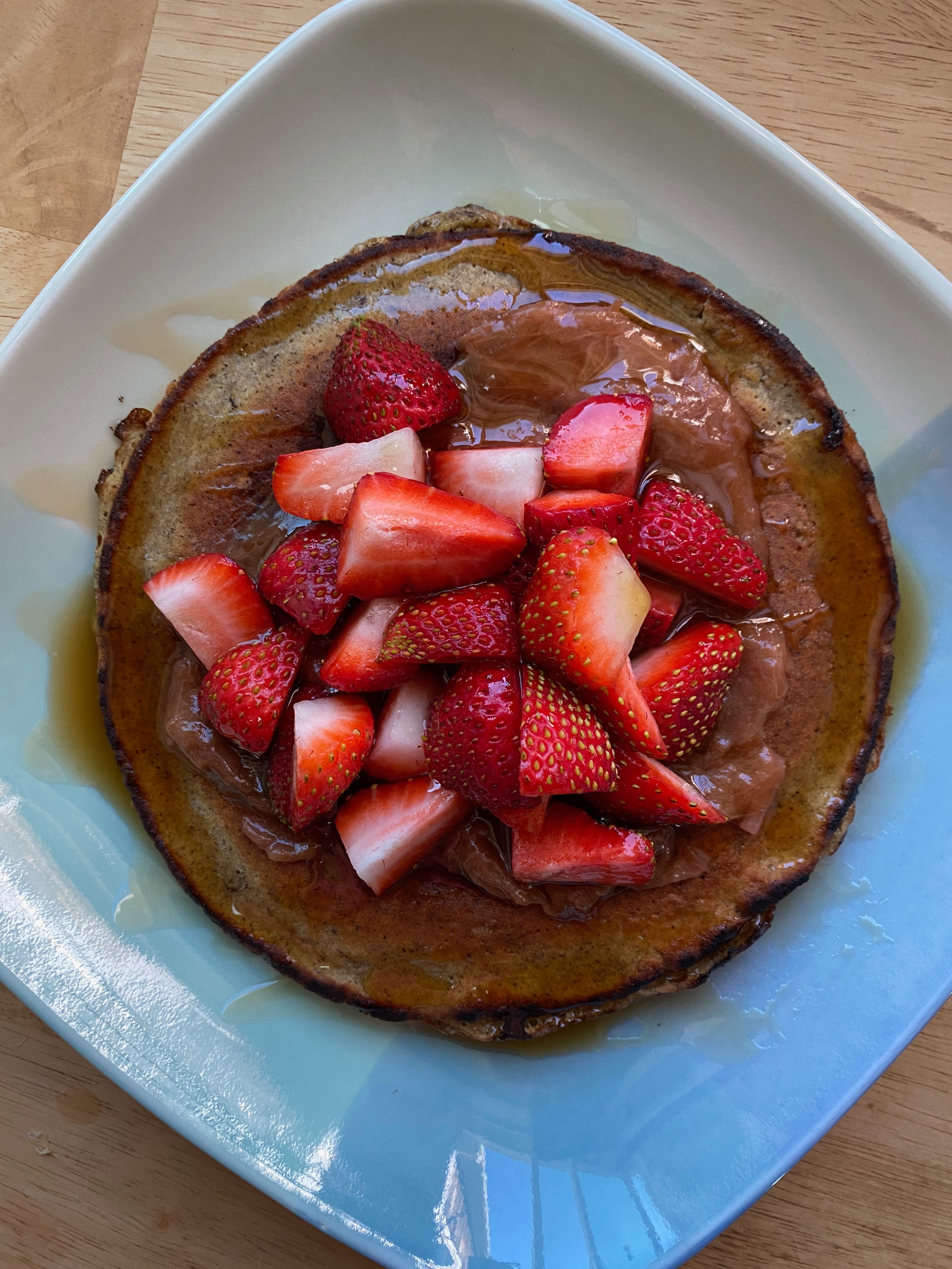 A large round buckwheat pancake sits on a blue and green square ceramic plate. The pancake is topped with a circle of rhubarb compote, a pile of sliced strawberries, and maple syrup.
