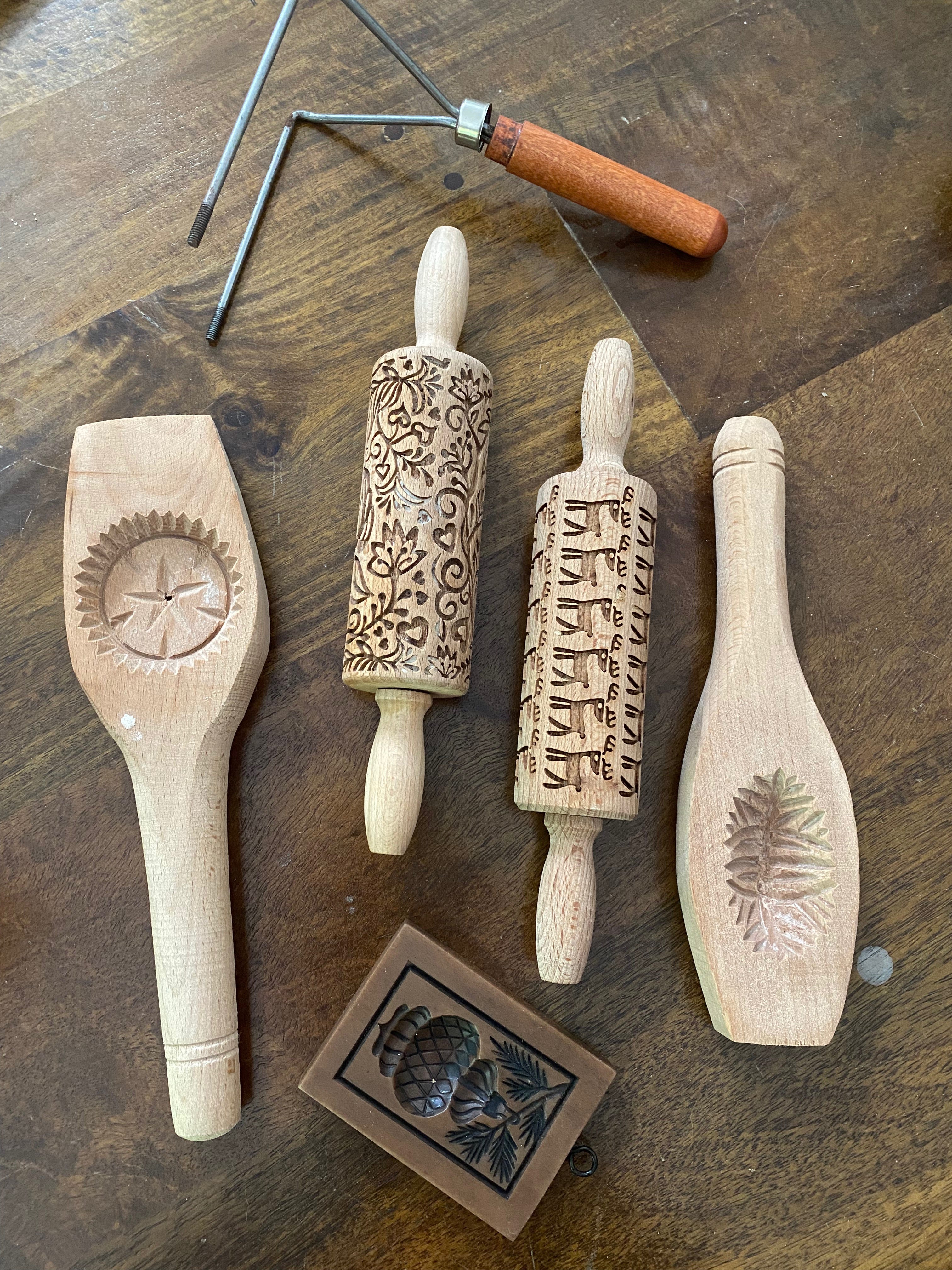 Several baking tools arranged on a wooden table. They include two ma’amoul molds, two small patterned rolling pins (one with a flower design and one with reindeer), a square springerle mold, and a small tool with a wooden handle and two metal prongs (part of a rosette iron).