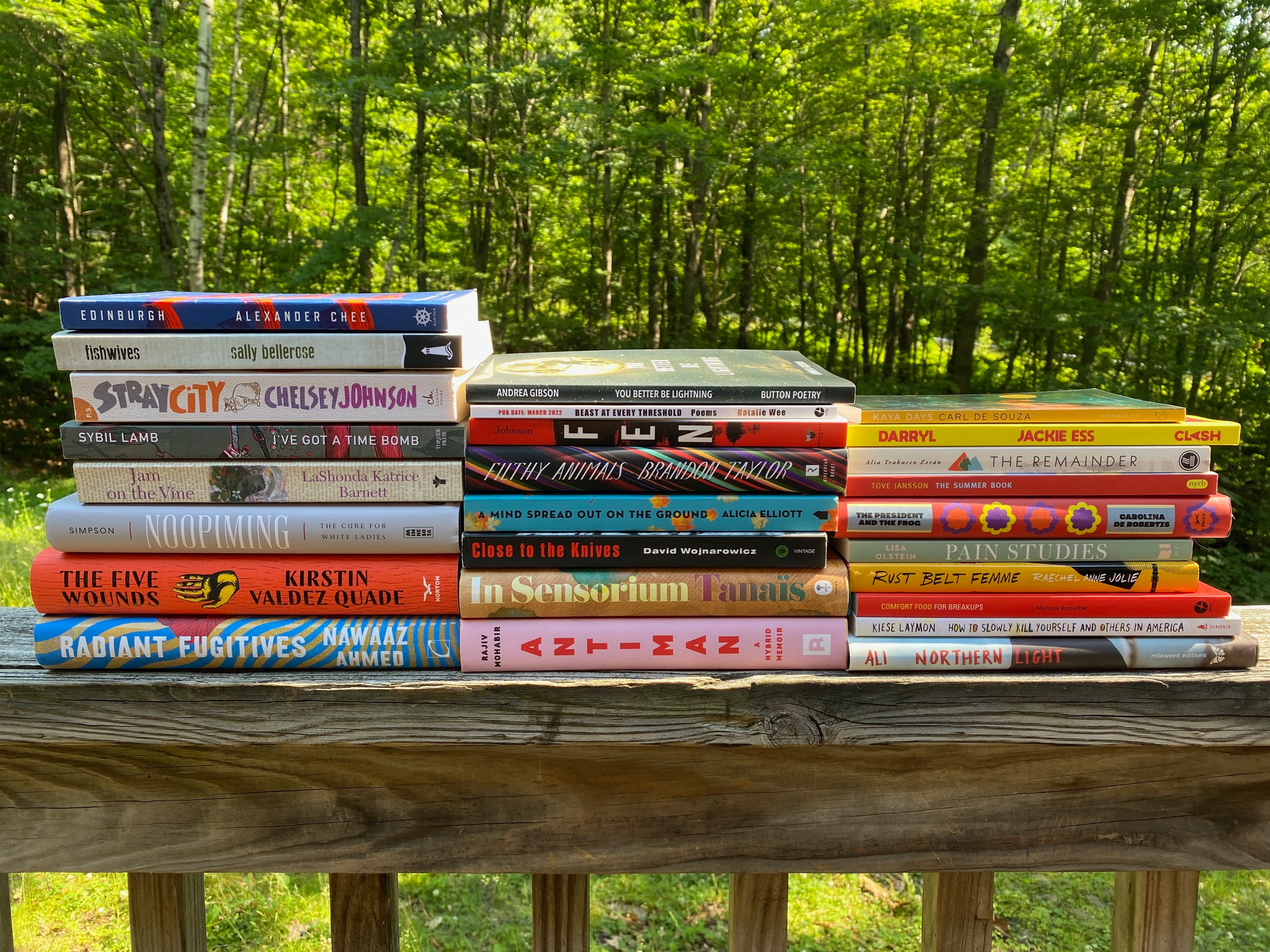 Three stacks of the books sit on a porch railing in front of sunlight trees. Books are:Radiant Fugitives, The Five Wounds, Noopiming, Jam on the Vine, I've Got a Time Bomb, Stray City, Fishwives, Edinburgh, Antiman, In Sensorium, Close to the Knives, A Mind Spread Out on the Ground, Filthy Animals, Fen, Beasts at Every Threshold, You Better Be Lightning, Northern Light, How to Slowly Kill Yourself and Others in America, Comfort Food for Breakups, Rust Belt Femme, Pain Studies, The President and the Frog, The Summer Book, The Remainder, Darryl, and Kaya Days.