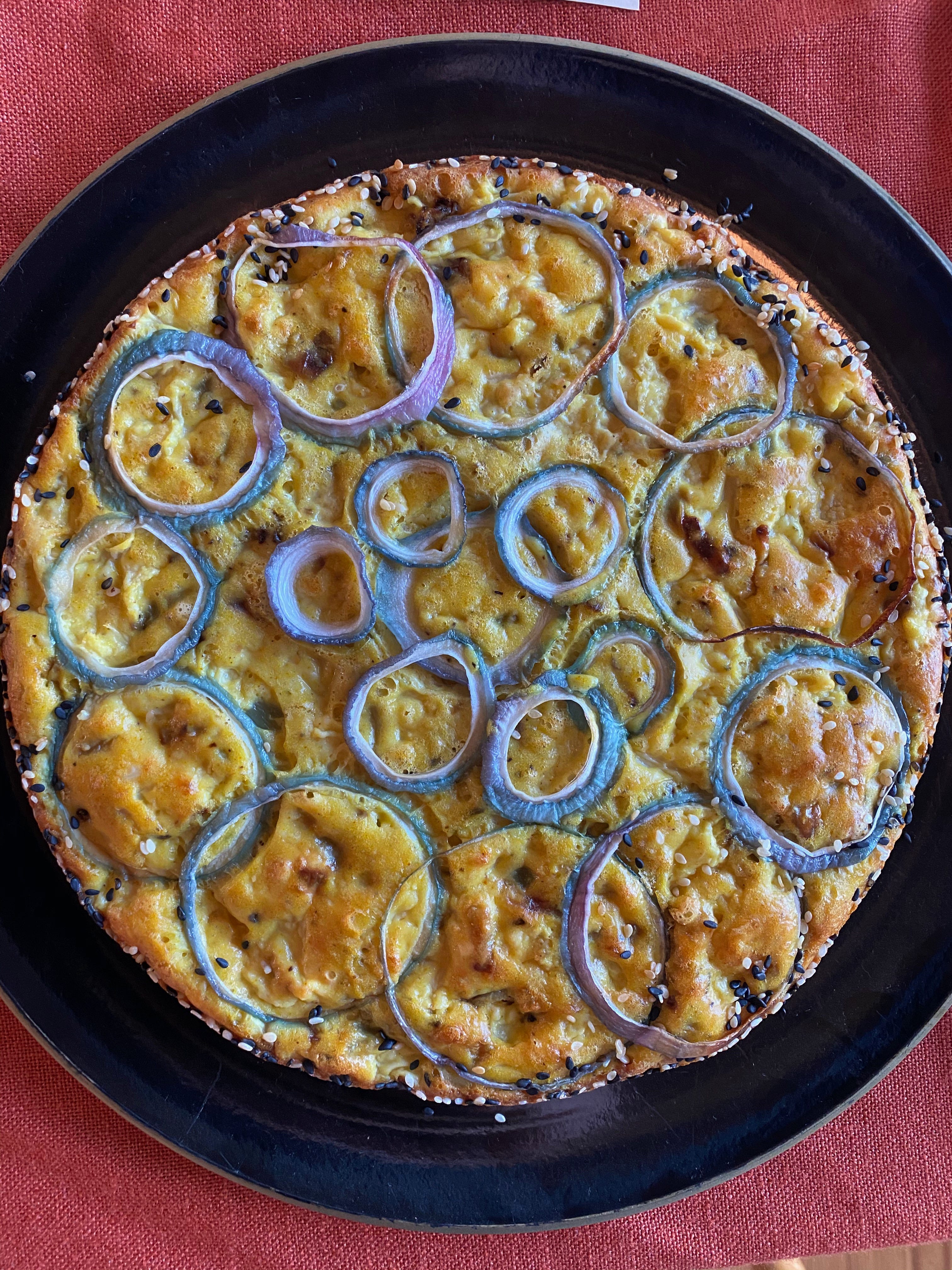 A golden brown eggy cake sits on a black platter. It is topped with rings of red onion, and the sides are coated in black and white sesame seeds.